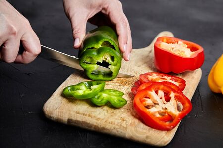 Pepper slicing. Hands, knife and red, green, yellow bell peppers, wooden cutting board on stone table. Sliced sweet peppers on black background. Vegetable ingredient, cooking diet salad, healthy foodの写真素材