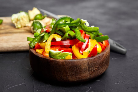 Chopped bell peppers in wooden bowl on black background. Red, green, yellow peppers. Colorful pieces...