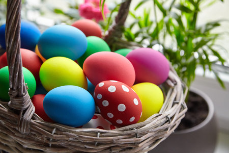 Basket with colorful Easter Eggs near window and blurred green leaves of houseplant on background....