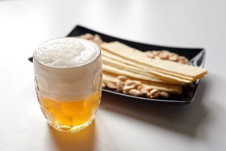 Mug of unfiltered light wheat beer with beer snacks on white table. White unfiltered beer, long...