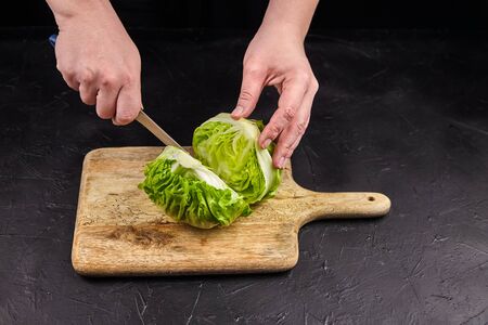 Iceberg lettuce (crisphead lettuce) on wooden cutting board. Person cuts iceberg lettuce on black stone table background. Leafy green vegetable, cooking salad, selective focusの写真素材