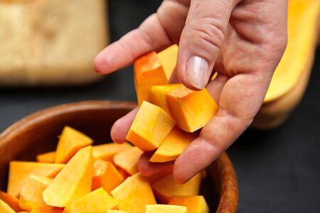 Pumpkin pieces in female hand. Chopped butternut squash in a wooden bowl on black background, closeup. Woman throws cutted pumpkin cubes into a salad dish. Cooking winter squash, food ingredientの写真素材