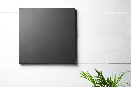 A black cotton canvas and leaves of green plant  on white wooden background. Stretched clean canvas hanging on wall. Mockup, front view