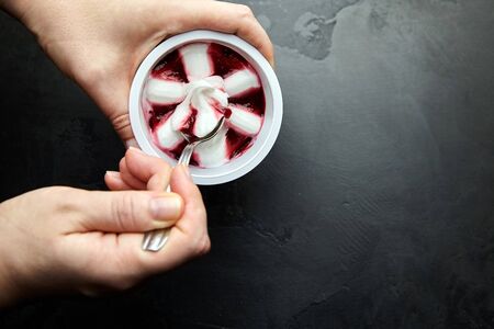 Yogurt ice cream in a white plastic cup on black background, frozen sweet fruit dessert. Spoon with a cherry icecream in female hand, closeup, top viewの写真素材