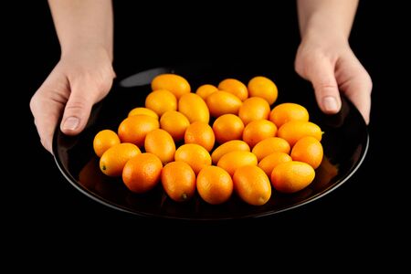 Kumquat, fresh citrus fruits, Nagami variety. Heap of cumquats in ceramic plate in hands on black background, selective focus. Eaten whole, healthy food, raw dietの写真素材