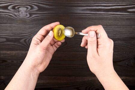 Eating kiwi gold fruit. Hands with spoon and half of ripe juicy kiwi with yellow flesh on wooden background, top viewの写真素材