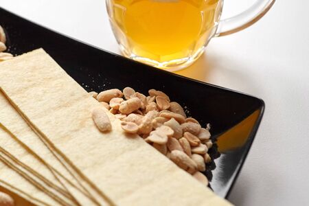Mug of unfiltered light wheat beer with beer snacks on white table. White unfiltered beer, long potato chips, salted roasted peeled peanuts in black plate, selective focusの写真素材