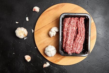 Ground beef in a plastic tray and white garlic cloves on wooden cutting board on black stone background, top view. Cooking food. Fresh raw minced meat and garlic headsの写真素材