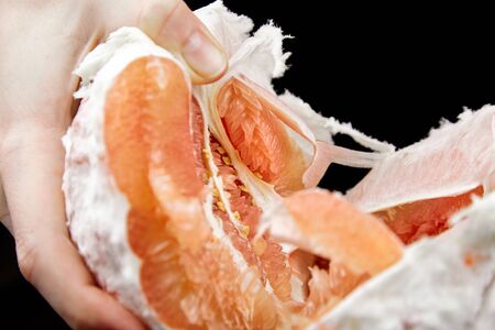 Pomelo citrus fruit with sweet red flesh on black background. Fresh juicy shaddock (Citrus maxima), closeup. Peeled pomelo in a female hand. Healthy fruitの写真素材