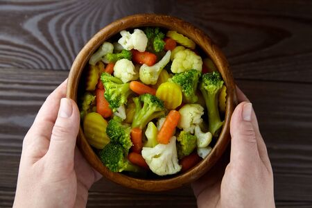 Vegetable mix. Vegan food. Boiled broccoli, cauliflower and carrot in wooden bowl in female hands on wooden background, top view. Healthy food. Steamed vegetablesの写真素材