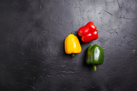 Red, green, and yellow bell peppers on black background with copy space. Three sweet peppers in different colors on stone table, vegetable ingredient, healthy food, top view, flat layの写真素材