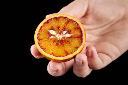 Blood or red orange fruit half with seeds and red juicy flesh in hand on black background. Person...