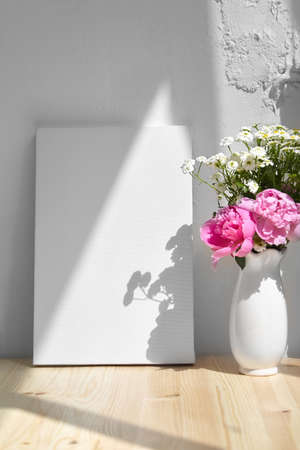 Mockup, vertical blank canvas, vase with pink flowers on wooden table, white wall with shadowの写真素材