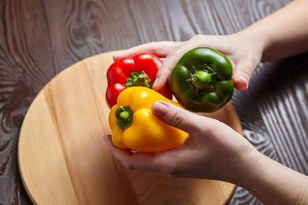 Red, green, and yellow bell peppers, cutting board on wooden background. Hands hold three sweet...
