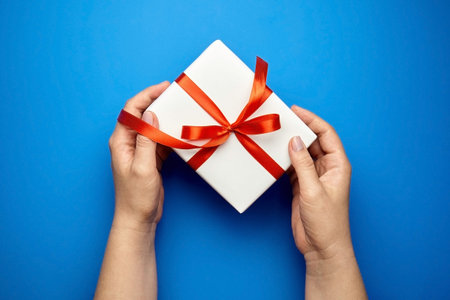 Hands with white gift box tied with red ribbon on blue background