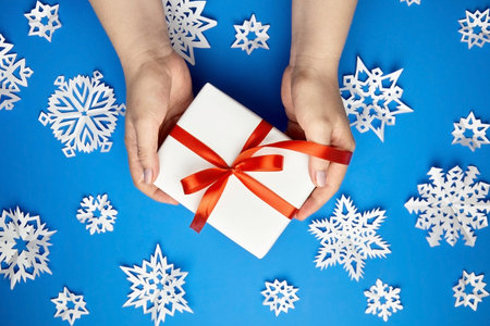 Hands holding white gift box with red ribbon on blue background with paper snowflakes