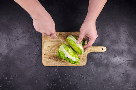 Iceberg lettuce (crisphead lettuce) on wooden cutting board, top view. Person cuts iceberg lettuce on black stone table background. Leafy green vegetable, cooking salad, selective focusの写真素材