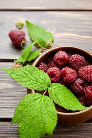 Fresh raspberry red berries with green leaves in bowl on wooden table background, ripe juicy...
