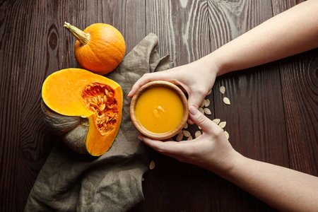 Female hands holding a bowl of pumpkin soup on dark wooden table background. Autumn thanksgiving day meals.の写真素材