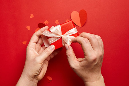 Hands holding valentines day gift box on red background with paper heartsの写真素材