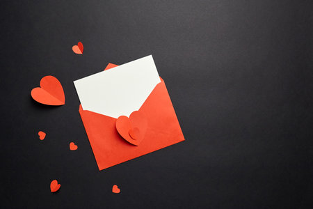 Greeting card mockup with paper decorations on black background, flat lay. Valentines Day blank greeting card or invitation, envelope and red paper heartsの写真素材