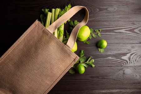 Reusable shopping bag with fresh fruits and greens on wooden background, top view. Jute bag (eco-friendly material), green apples, limes, celery on brown table. Healthy food, useful organic productsの写真素材