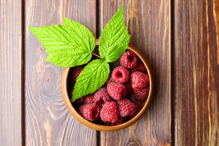 Fresh raspberry red berries with green leaves in bowl on wooden table background, ripe juicy...