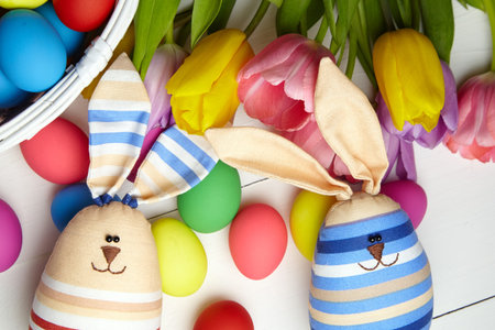 Easter bunny doll and Easter eggs and spring colorful flowers on white wooden table. Painted chicken...