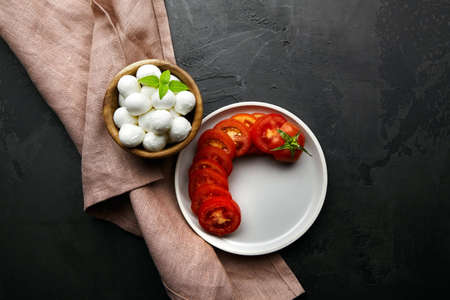 Mini mozzarella cheese balls in wooden bowl and fresh ripe tomatoes on linen napkin over dark stony background, top viewの写真素材