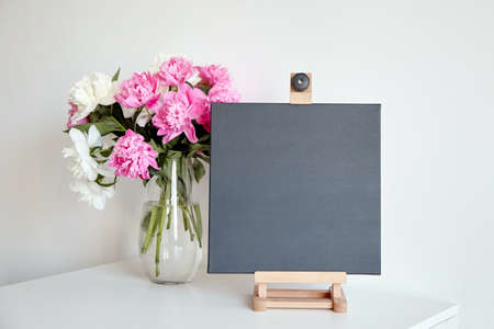 Black canvas mockup on wooden easel and vase with pink flowers on table on white wall background. Blank artistic canvasの写真素材