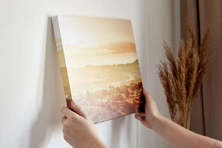 Canvas print with gallery wrap and dry grass interior decor. Woman hangs landscape photography on white wallの写真素材