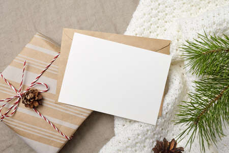 Christmas greeting card mockup with envelope, gift box and pine tree branch with cones on knitted backgroundの写真素材