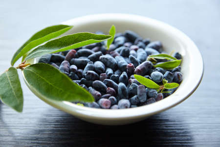 Haskap berry with fresh green leaves in white bowl on wooden table. Honeyberry (Lonicera caerulea) or honeysuckle on wood backgroundの写真素材