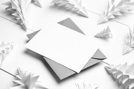 Holiday card mockup with envelope and white paper fir trees on white wooden background. Christmas...