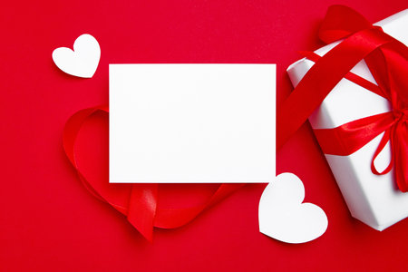 Valentine Day card mockup with white hearts and gift box on red background, top view, flat lay. Blank invitation, flyer, greeting card mockup with holiday decor. Empty love letter with copy spaceの写真素材