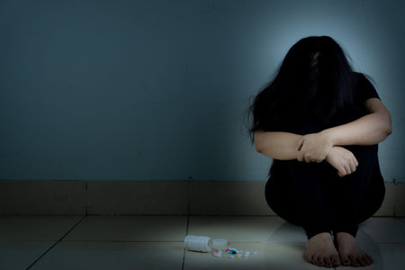 sad woman hug her knee and cry sitting alone with assorted pharmaceutical medicine pills in a dark...