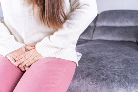 Woman hands holding her crotch suffering from painful stomachache while sitting on sofa at home....