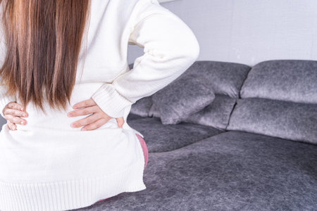 Young woman suffering from back pain while sitting on sofa at home. Healthcare medical or daily life...