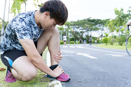 Asian man use hands hold on his ankle while running on road in the park. Injury from workout concept.の写真素材