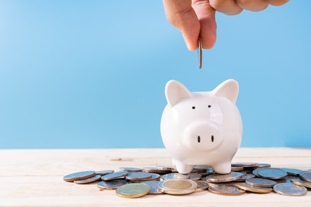 Hand putting money coin into piggy bank with a pile coins isolated blue background. Saving money and investment concept.の写真素材