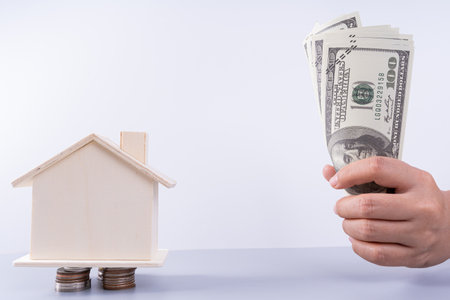 Hand holding money paper and wooden house on stack coins isolated grey background. Property investment and house mortgage financial concept.の写真素材