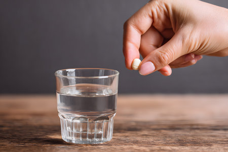 A hand holds a small pill above a glass of clear water.の素材