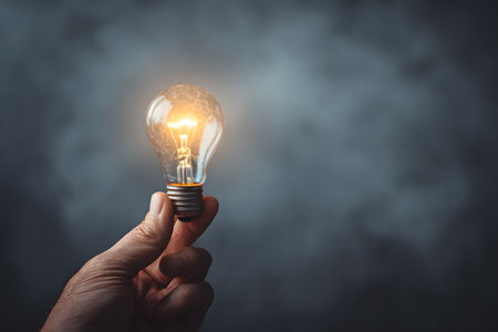 A glowing light bulb is held in a hand against a dark, smoky background.の素材