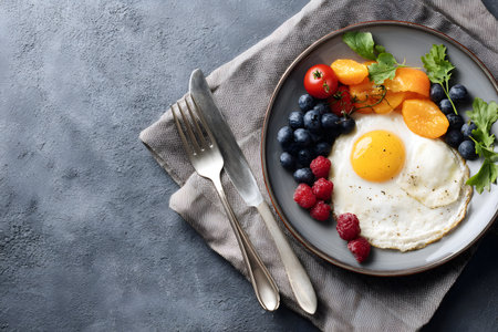 A delicious breakfast plate with a fried egg, berries, and fruits rests on a textured surface.の素材