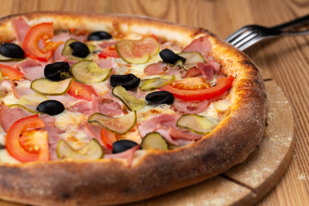 hearty italian pizza on a wooden board. pizza with ham, tomatoes, pickles and olivesの写真素材