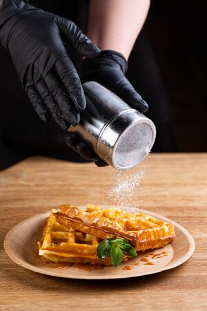 Viennese waffles on a disposable plate with caramel syrup, icing sugar and mint. chef decorates dessert with powdered sugarの写真素材