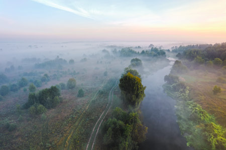 Aerial panoramic view of misty countryside in the summer morning hour with river, meadows, trees.