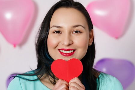 Valentine's Day. Beautiful young woman with heart in her hands. Young woman with red heart on white background with inflatable balls. portrait of attractive smiling woman on white studio shot with hearts. close upの写真素材