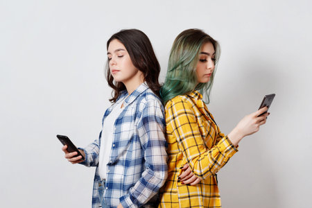 Modern technologies and internet addiction concept.Two young womans standing back to back, absorbed...