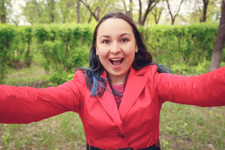 close-up photo. A young woman in red clothes on the street takes a selfie, looks at the camera and smiles. daylight in the Parkの写真素材
